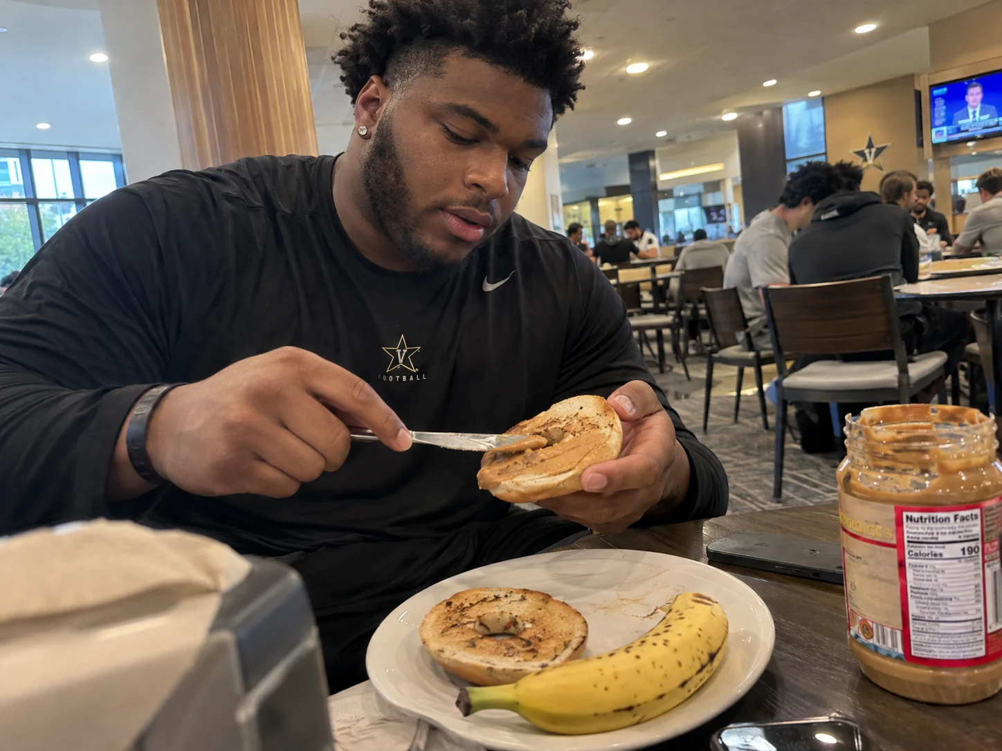 Football player spreading peanut butter on a toasted bagel beside a banana in the team dining room