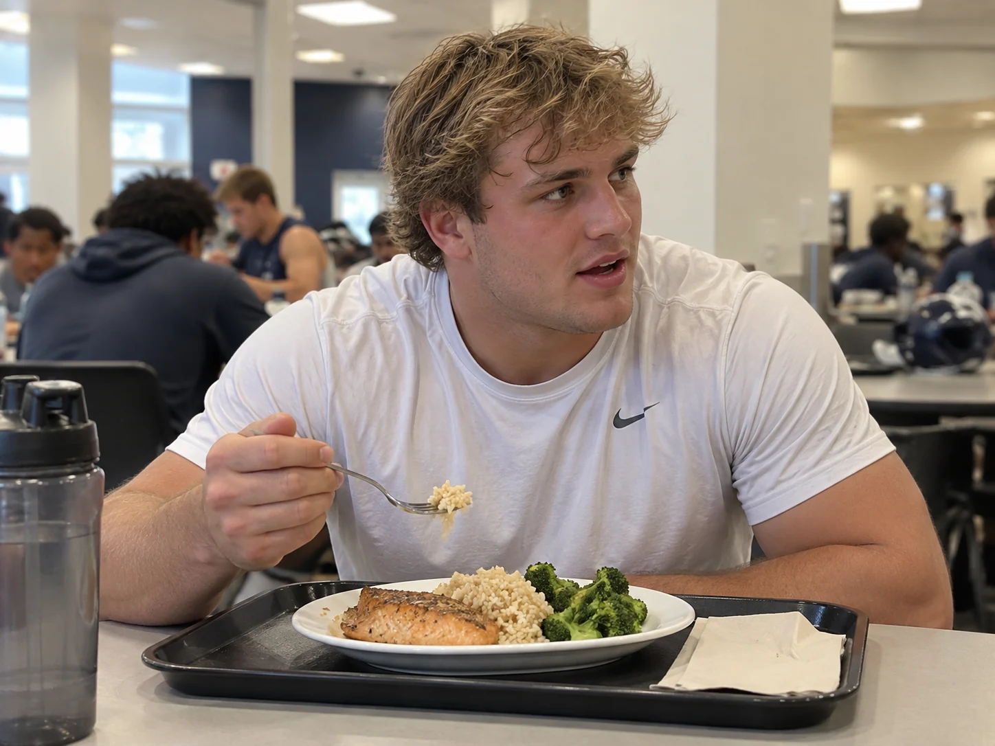 College football player seated with a plate of salmon, rice, and broccoli in a team dining area